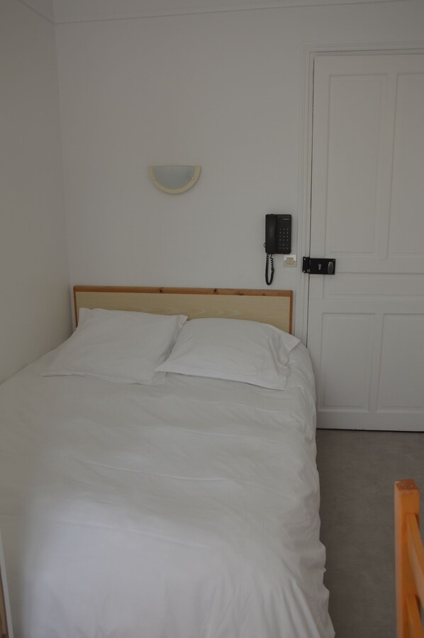 Una cama bien hecha con sábanas blancas en una habitación con cabecera de madera. Se ve un teléfono de pared y un interruptor de luz.