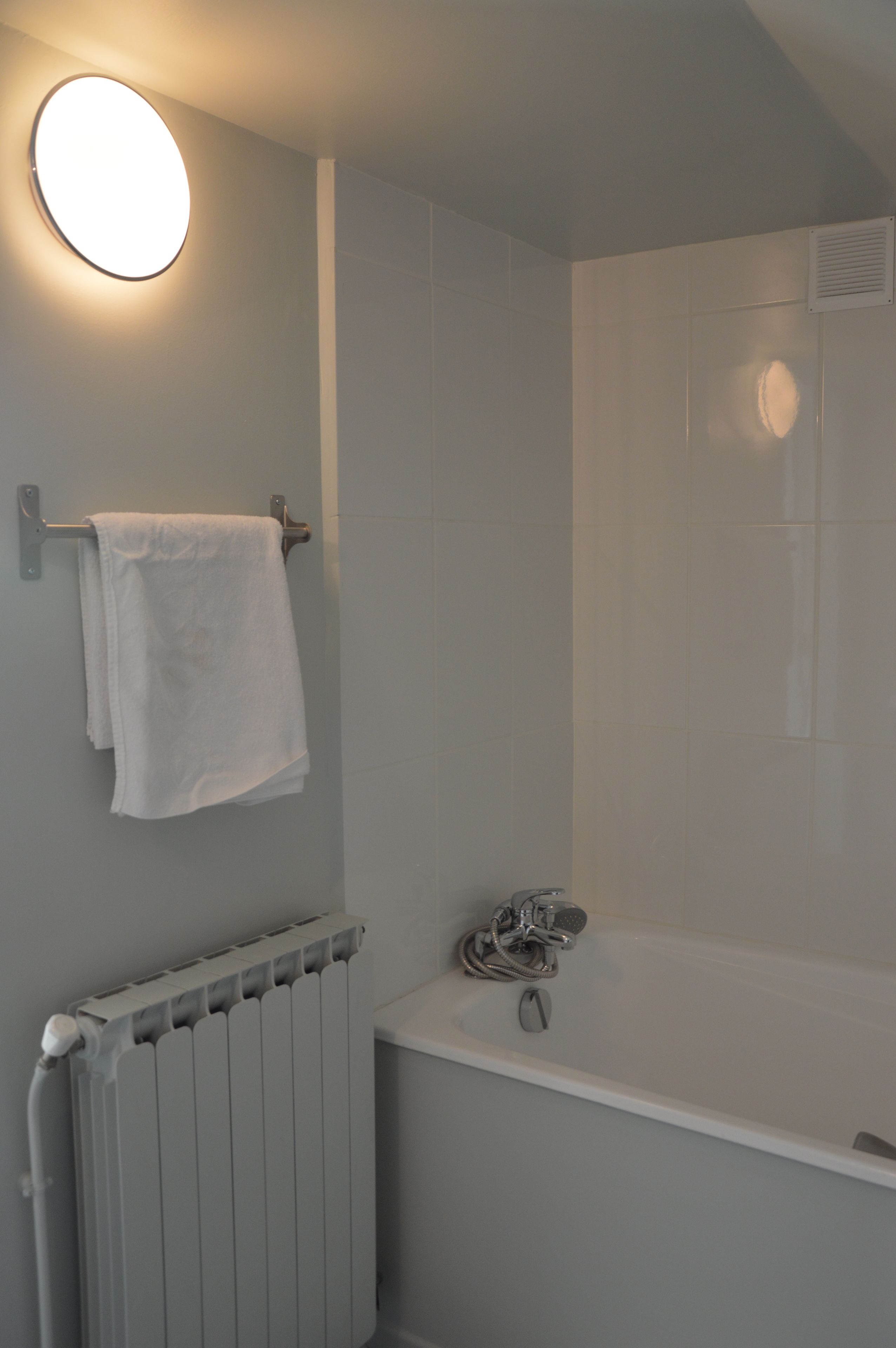 A bathroom with a white bathtub, a towel hanging on a metal rack, and a round light fixture on the wall.