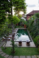 A swimming pool with lounge chairs, surrounded by greenery and a house in the background.