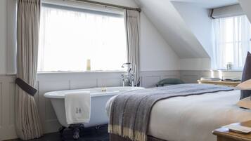 A bedroom with a large bed, a window with curtains, a bathtub, and a bedside table.
