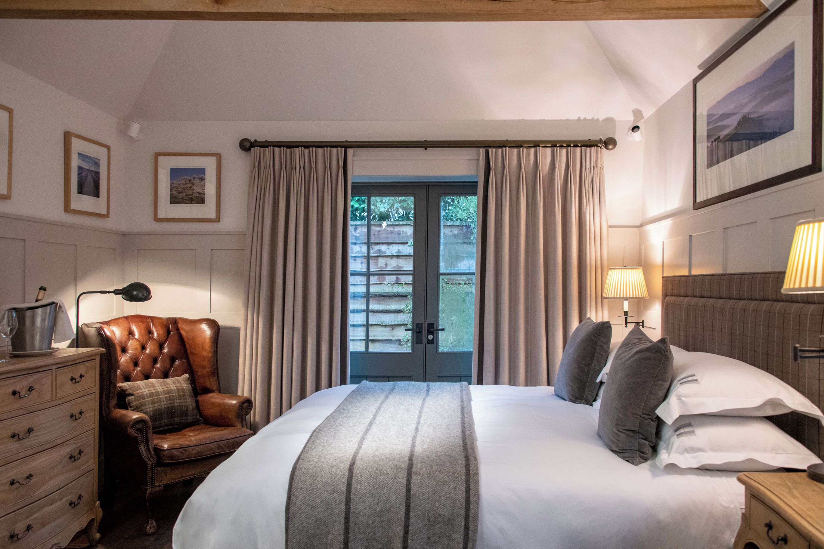 A bedroom with a large bed, a leather armchair, a wooden dresser, and a view through a door.