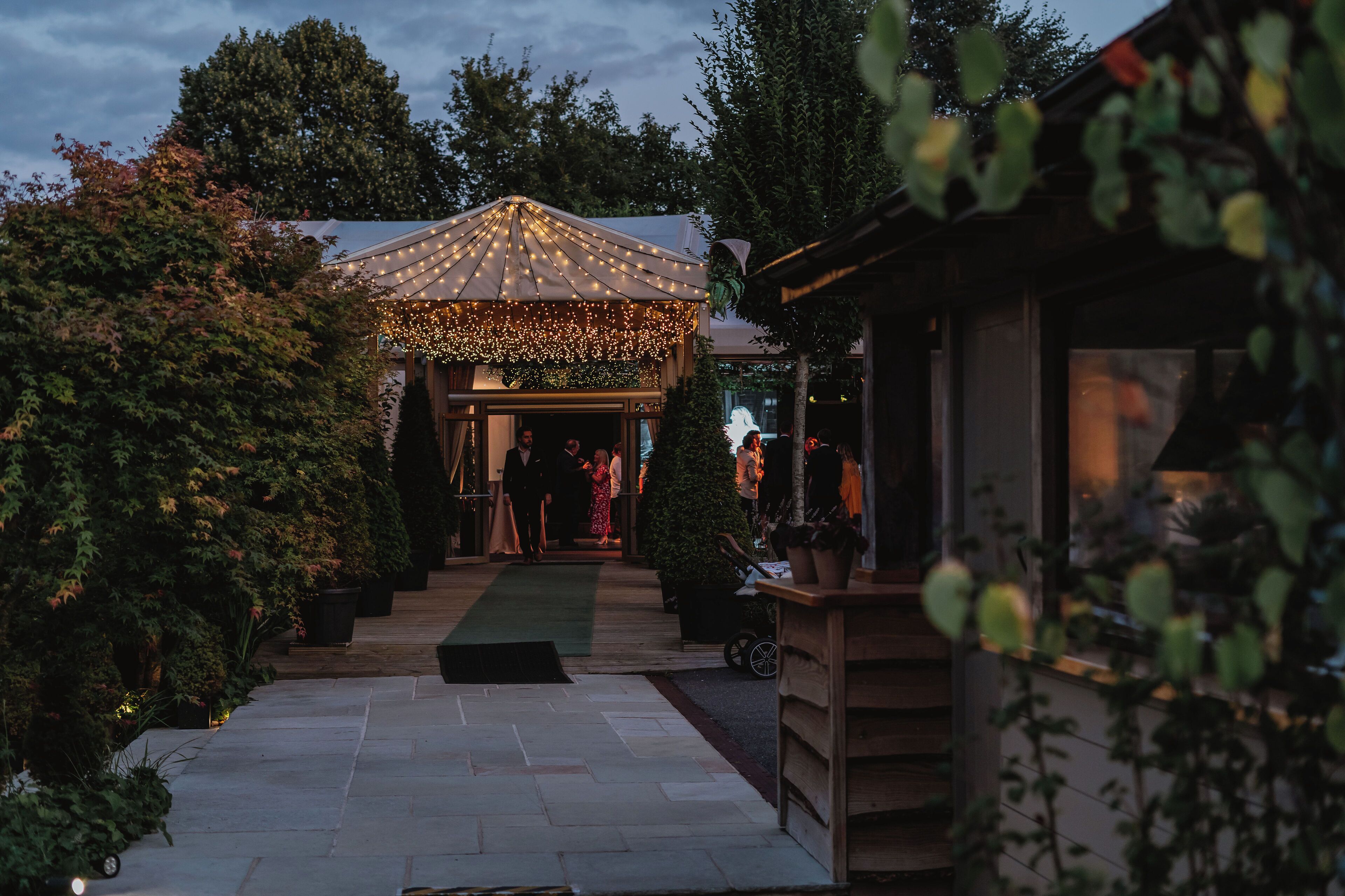 A covered outdoor area with a pathway leading to a building, surrounded by greenery.