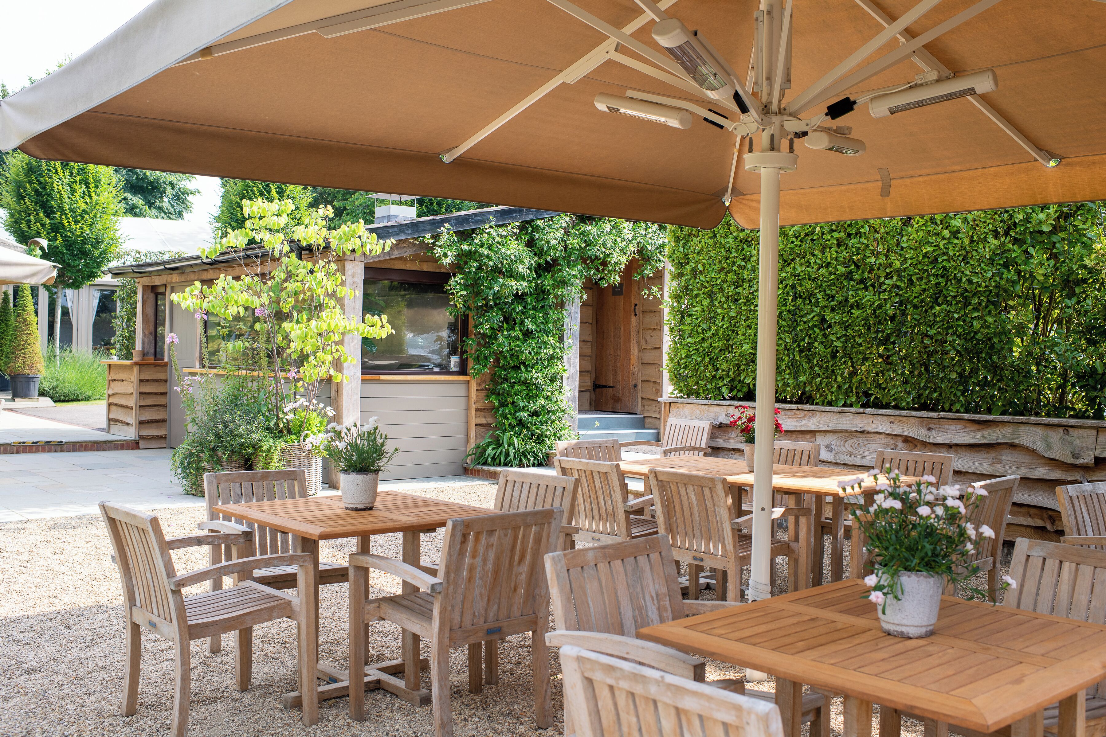 Une terrasse extérieure avec des tables et des chaises en bois, un grand parasol et des plantes en pot.