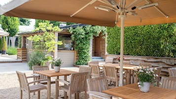 An outdoor patio with wooden tables and chairs, a large umbrella, and potted plants.
