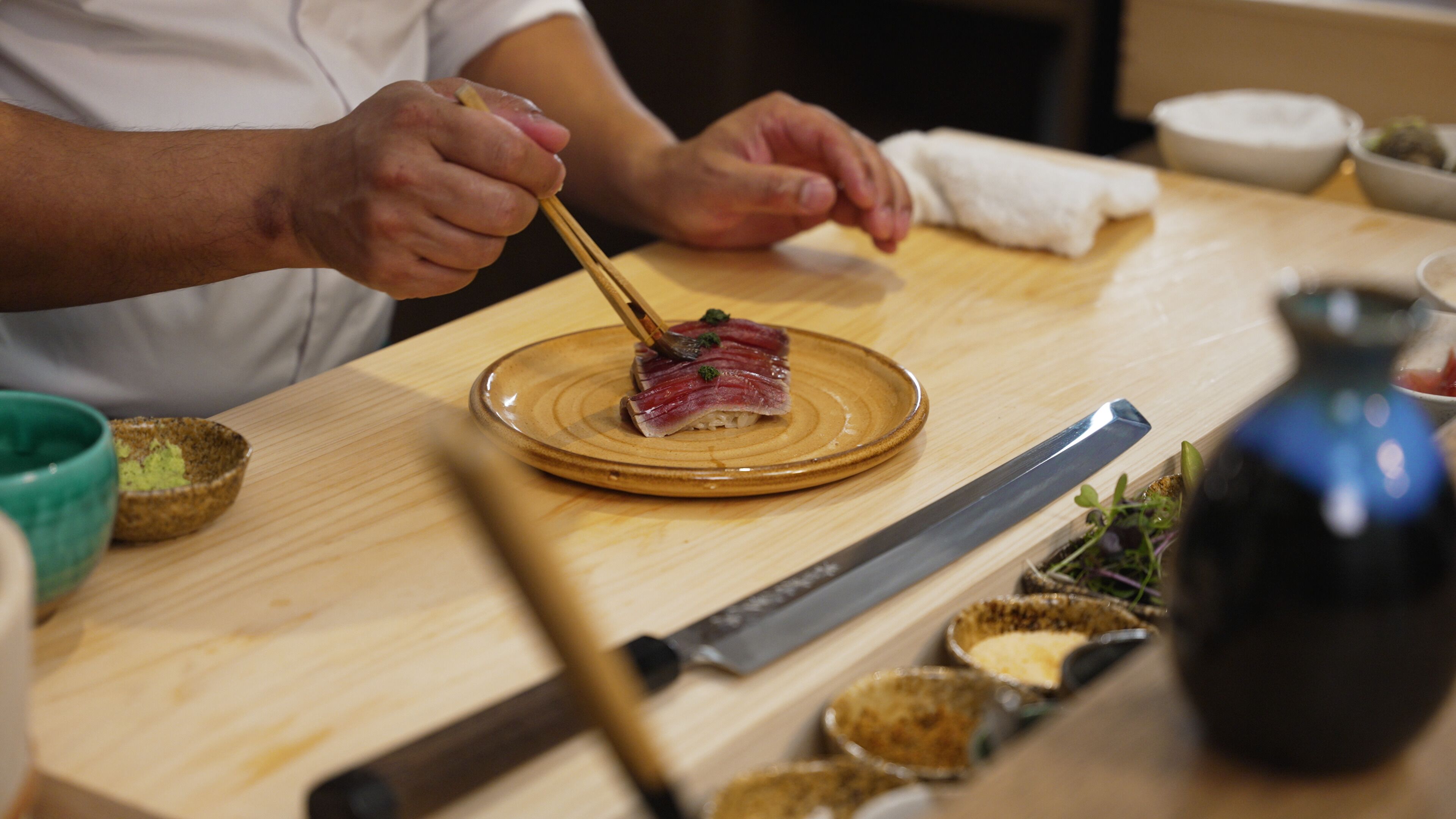 A wooden table with a plate of sashimi, chopsticks, a knife, and small bowls of condiments.