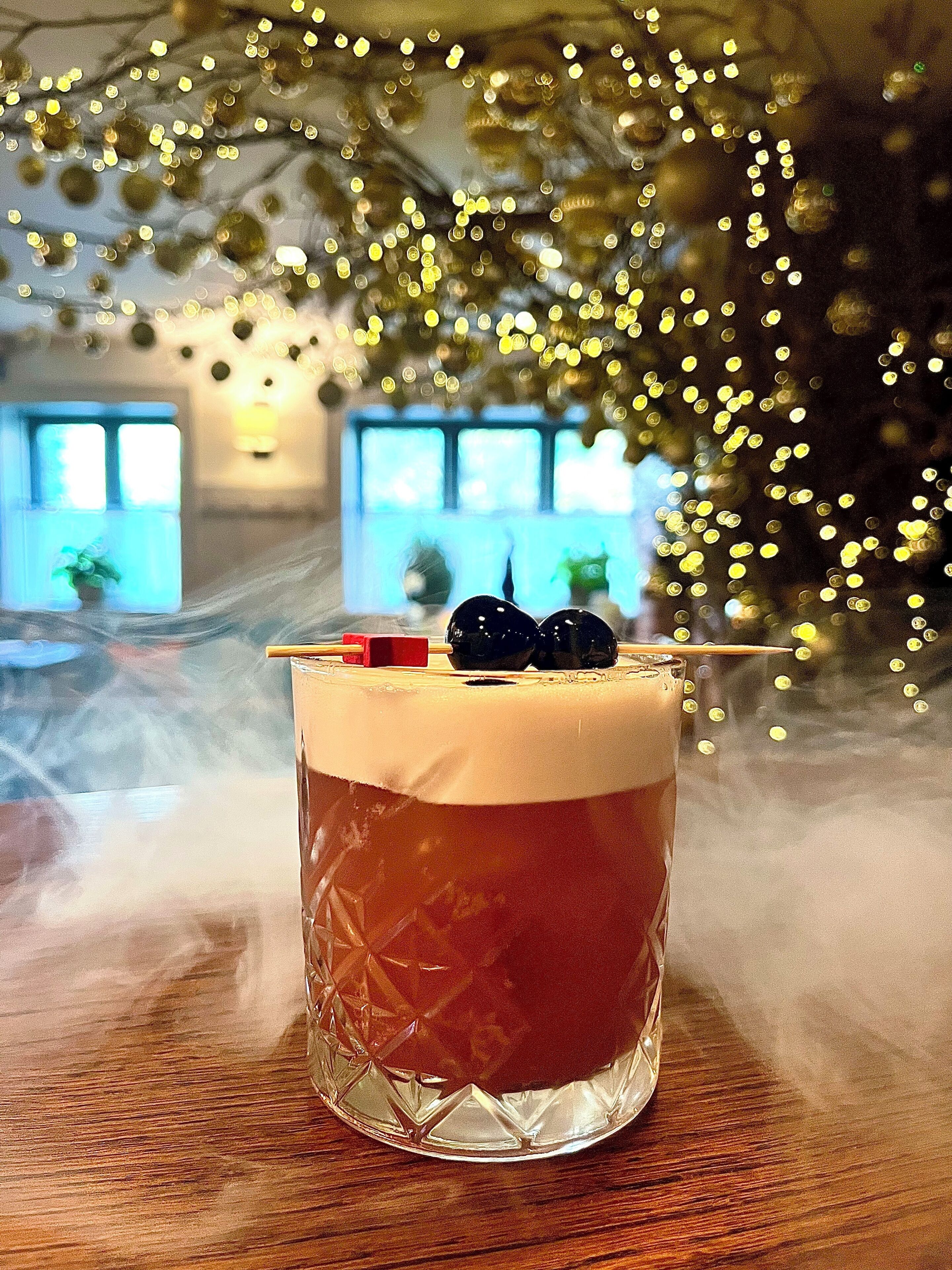 A cocktail in a glass with a straw and a cherry on top, placed on a wooden table with a blurred background of windows and lights.