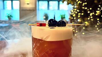 A cocktail in a glass with a straw and a cherry on top, placed on a wooden table with a blurred background of windows and lights.