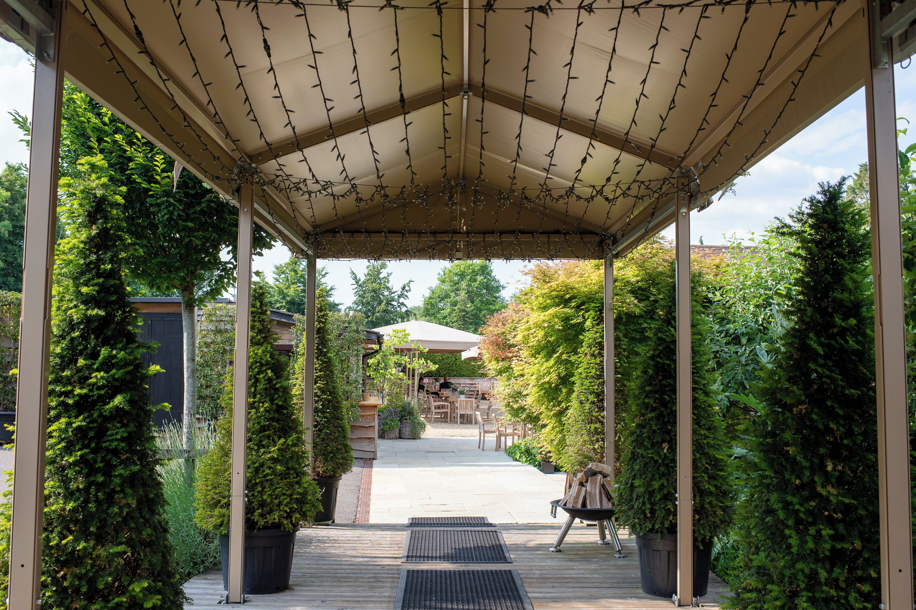 A covered patio area with a pergola structure, surrounded by lush greenery and a view of a backyard with outdoor furniture.