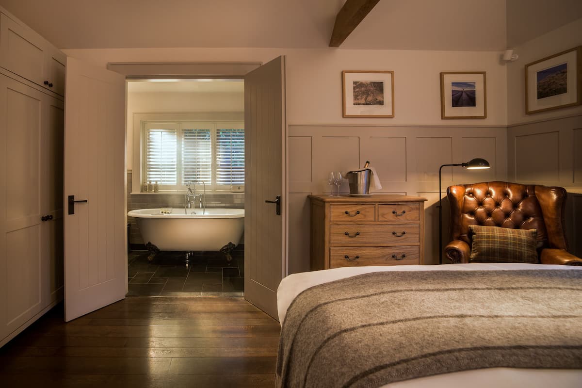 A bedroom with a large bed, a wooden dresser, a leather armchair, and a view of the bathroom with a bathtub and window.
