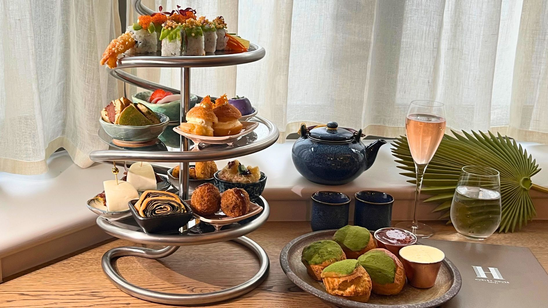 A tiered tray with various pastries and desserts, a teapot, and a glass of champagne on a table with a view of a window and a plant.