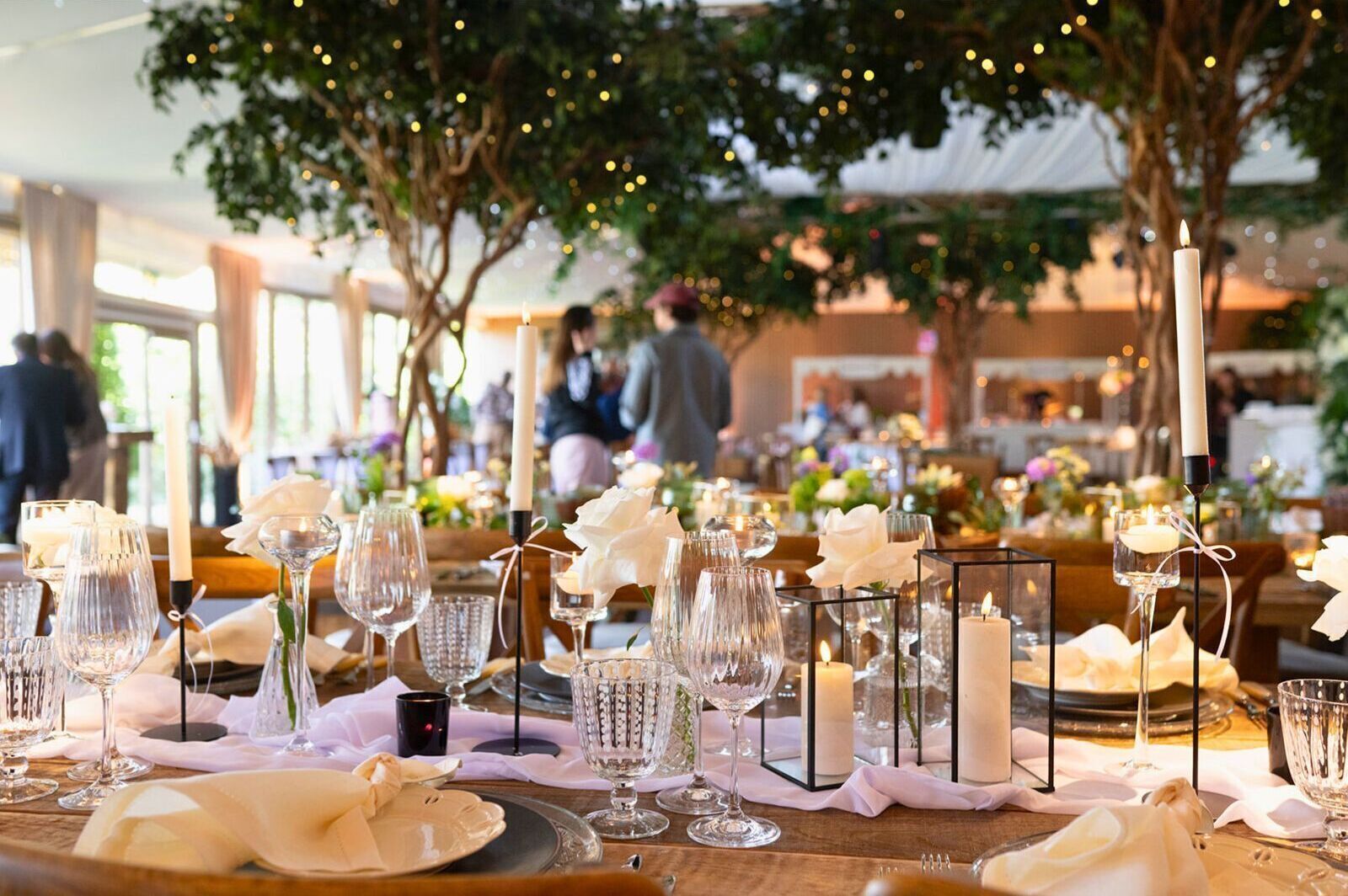 A well-set dining table with candles, glassware, and white napkins, under a tree with string lights.