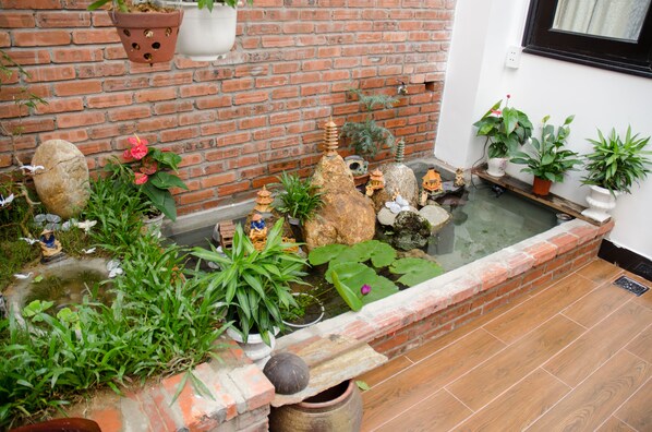 Un pequeño estanque con rocas y plantas, un muro de ladrillo y una ventana con cortina.