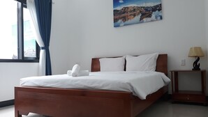 Una cama bien hecha con sábanas blancas y una toalla enrollada, un cabecero de madera, una mesita de noche con una lámpara, y un cuadro de un pueblo costero en la pared.