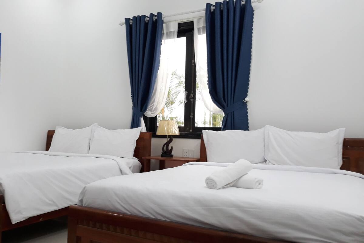 Une chambre d’hôtel avec deux lits simples, une table de chevet, une lampe et une fenêtre avec des rideaux bleus.