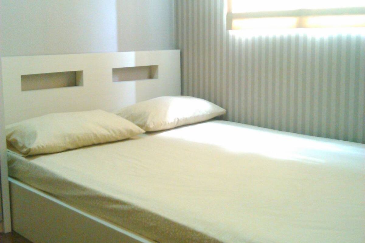 Een eenpersoonsbed met hoofdbord, een raam met zonwering en een effen muur.