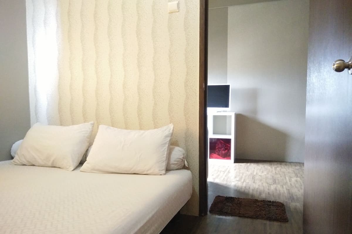 Een slaapkamer met een bed, kussens, een nachtkastje, een aan de muur bevestigde televisie en een deur naar een andere kamer.