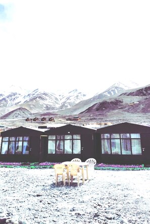 A hotel with multiple rooms, each with large windows and a view of snow-capped mountains. There are outdoor seating areas with chairs. - TIH Pangong Delight Camps and Cottages (Leh)