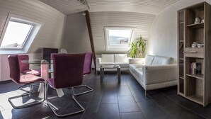 Un loft moderno con techo inclinado, una mesa de comedor de vidrio, sillas moradas, un sofá blanco, una estantería de madera y vistas a través de un tragaluz.
