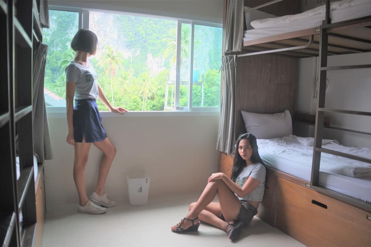 A dormitory room with bunk beds, a window with a view of greenery, and a white wall.