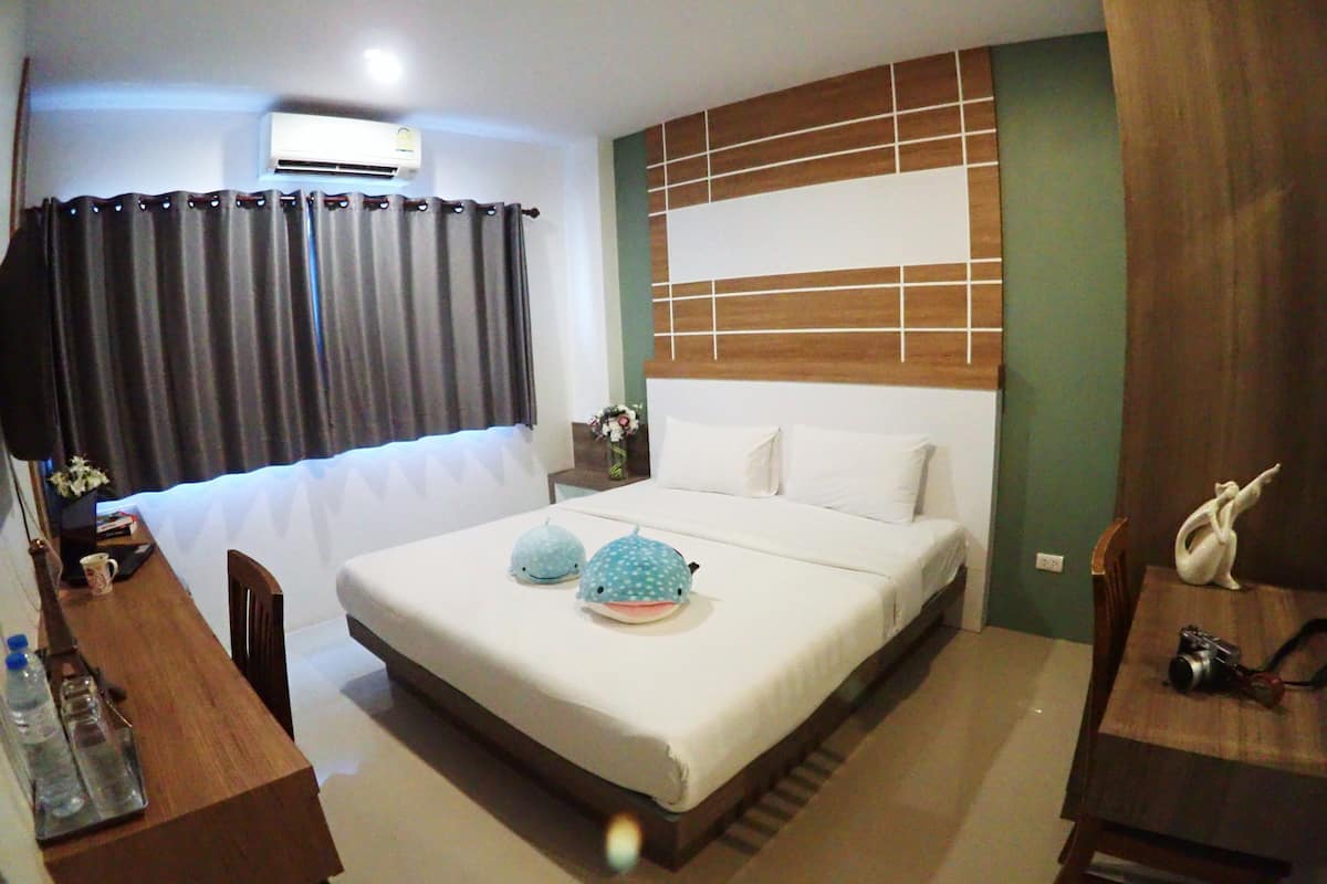 A hotel room with a bed, a desk, a chair, an air conditioner, a window with curtains, and a wall with a wooden panel design.
