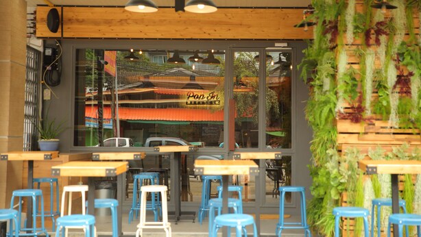 An indoor cafe with wooden tables and blue chairs, large windows, and hanging plants. - Pop-In Hostel Aonang (Krabi)