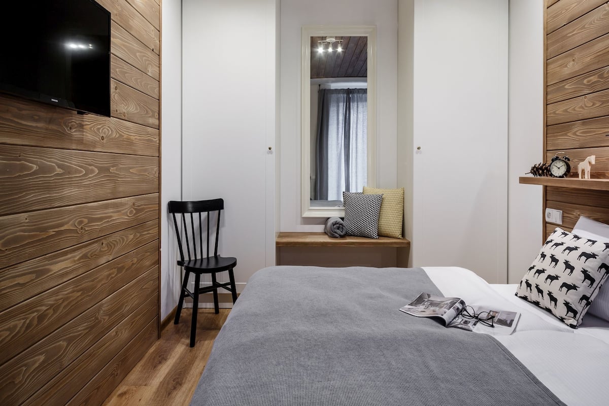 Una habitación compacta con cama, una silla, televisión montada en la pared y una ventana con cortinas.