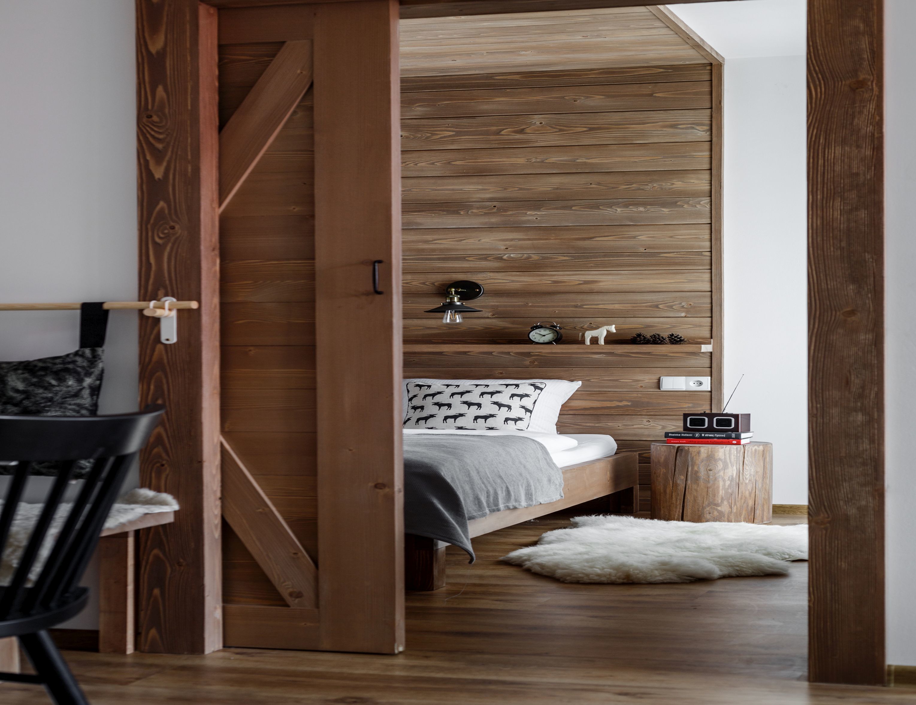 A bedroom with a wooden slat wall, a bed with patterned pillows, a small bedside table with a radio, and a fluffy rug.