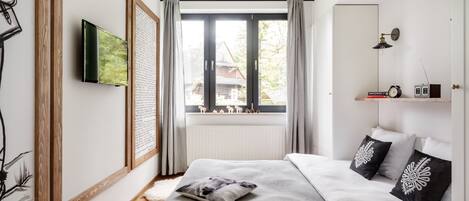 Quarto moderno com uma cama grande, televisão, janela com cortinas e mesa de cabeceira com abajur e livros.