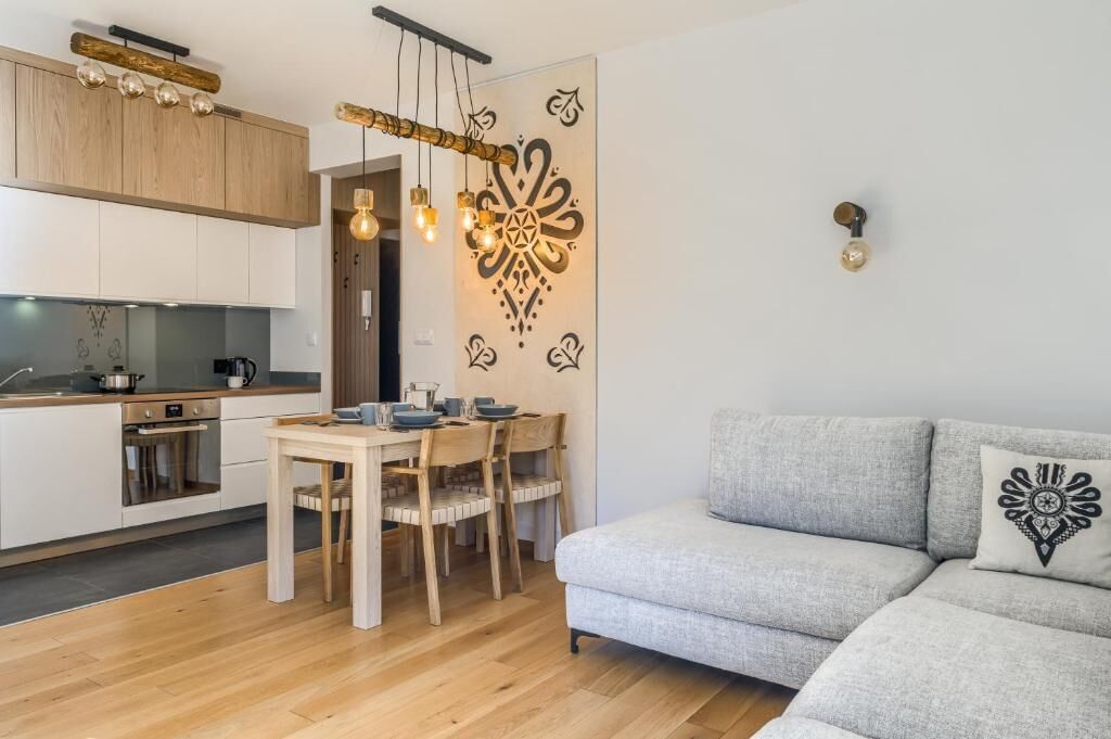 A modern kitchen with wooden cabinets, a dining table with chairs, a grey sofa, and a decorative wall piece.