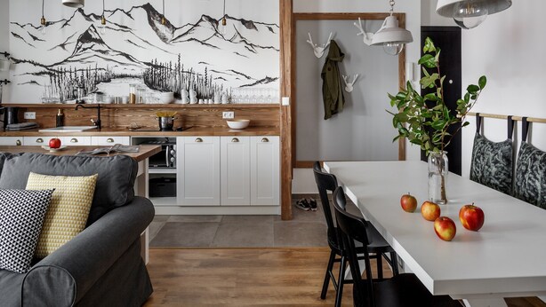 A modern kitchen with wooden cabinets, a white countertop, and a mountain landscape mural. There is a dining table with chairs and a vase with apples. - Tatrytop Apartamenty Granitica (Zakopane)