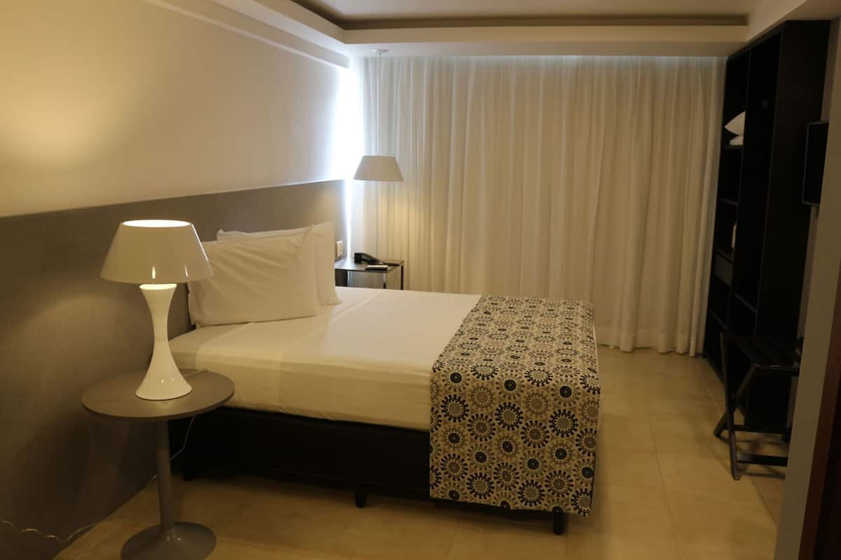 Habitación de hotel con cama, lámpara de noche, mesita de noche, manta decorativa, armario y ventana con cortinas.