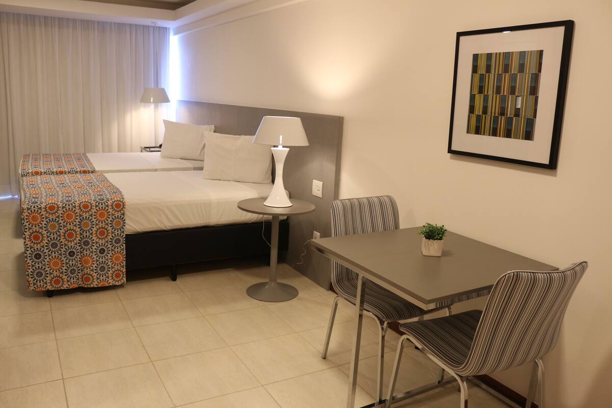 Una habitación de hotel con cama, mesita de noche, lámpara, silla y una pequeña mesa con una planta.