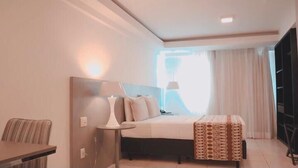 A hotel room with a bed, bedside table, lamp, and a view of the outdoors.
