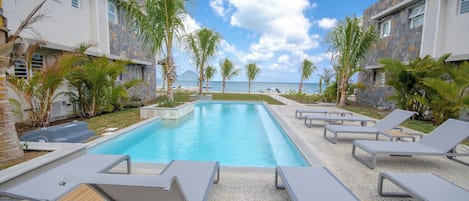 Área de piscina com espreguiçadeiras e palmeiras, com vista para um corpo de água.