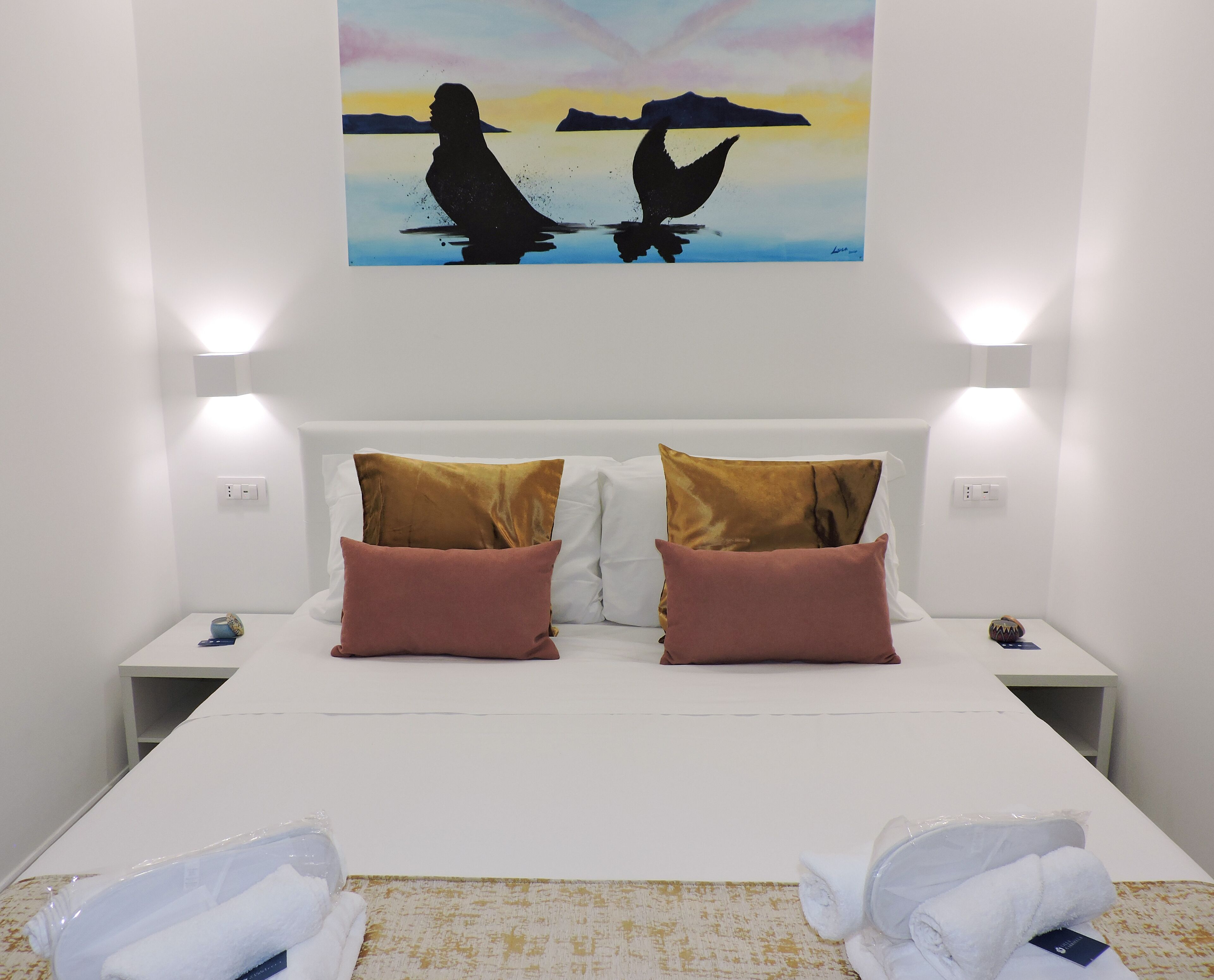 Quarto de hotel com cama, duas almofadas, um quadro e mesa de cabeceira com abajur.