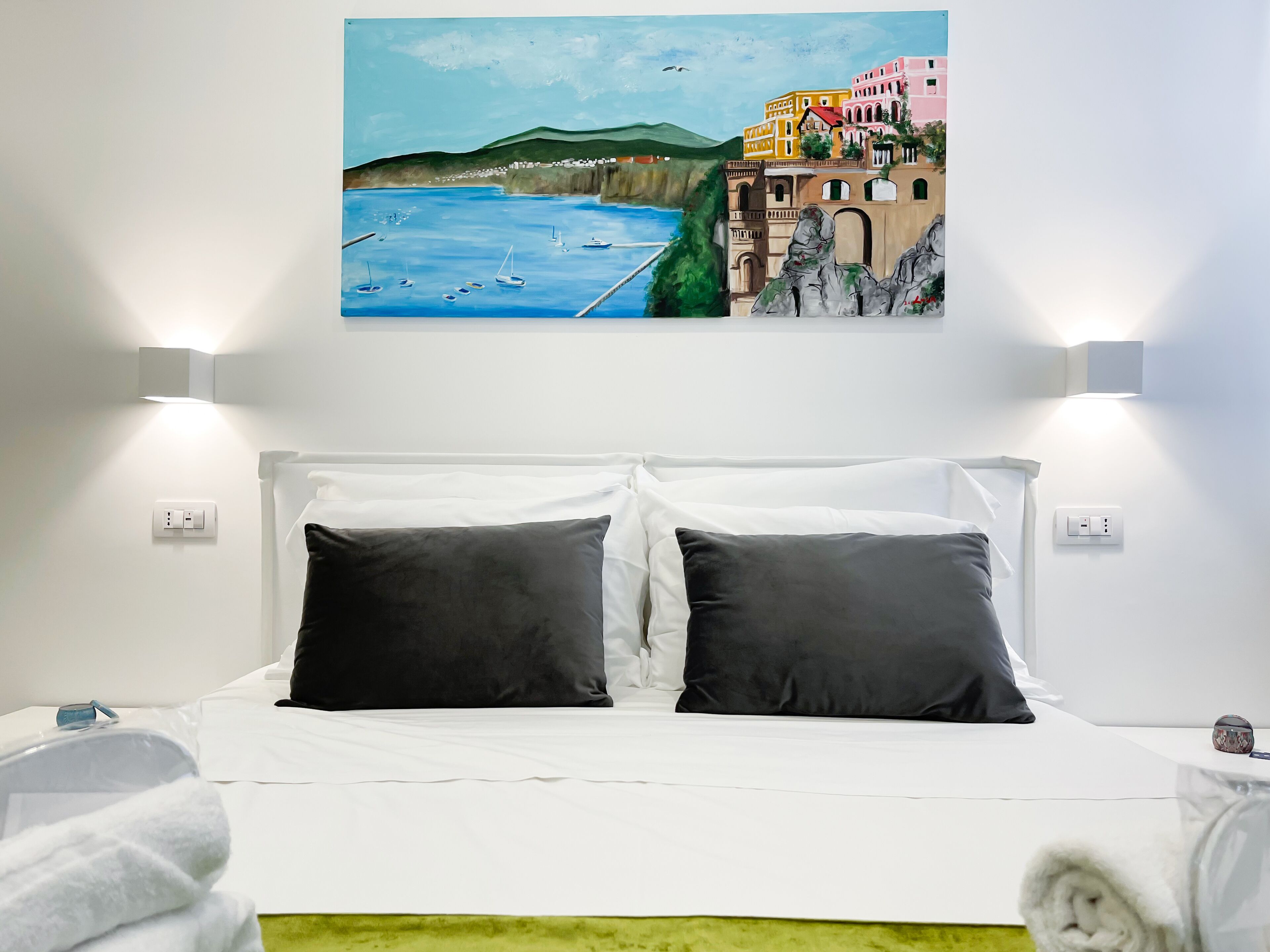 Habitación de hotel con cama, ropa de cama blanca, dos almohadas negras, un cuadro de una escena costera y un tapete verde.