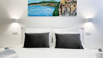 Habitación de hotel con cama, ropa de cama blanca, dos almohadas negras, un cuadro de una escena costera y un tapete verde.