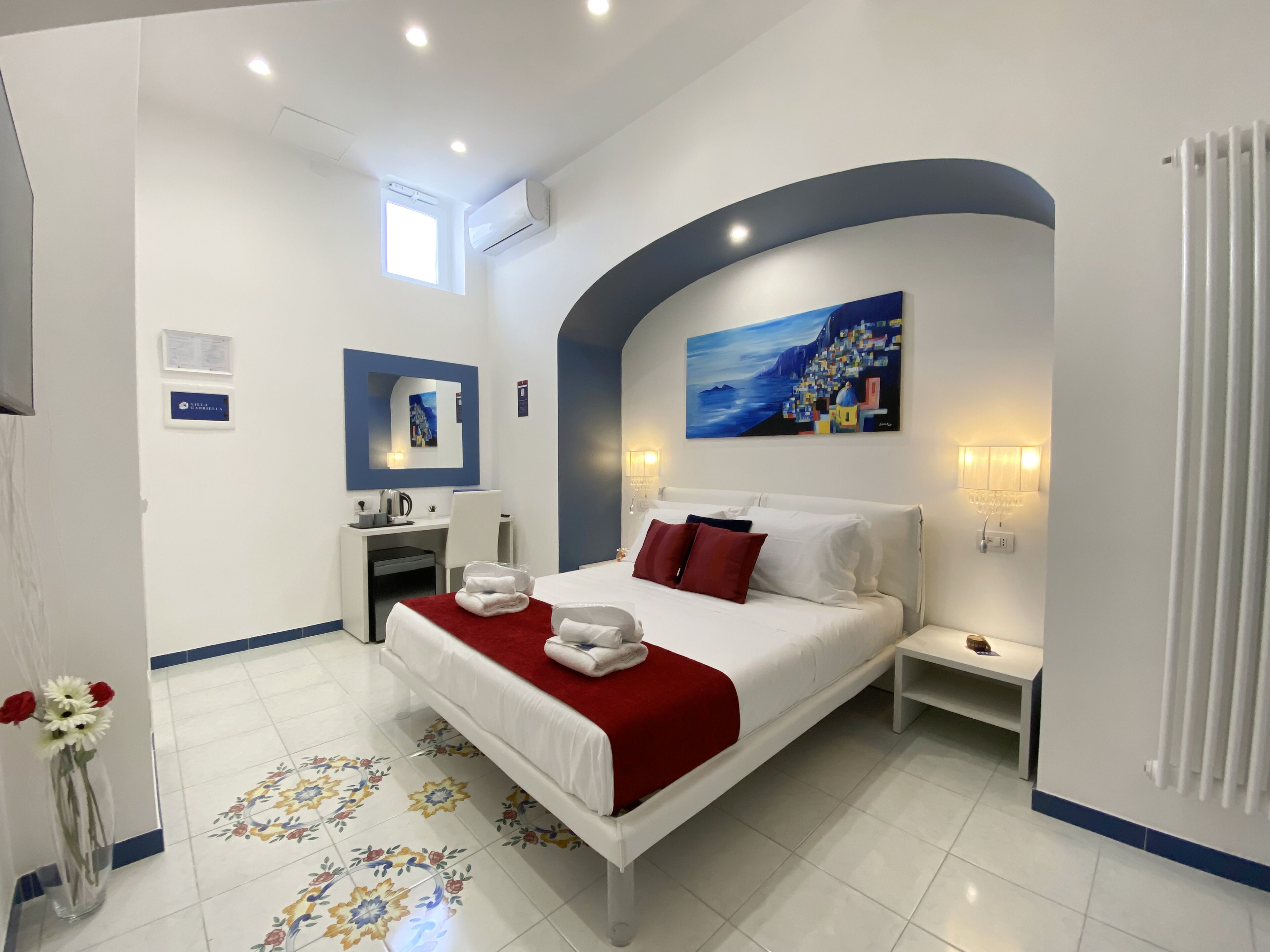 Een moderne hotelkamer met een groot bed, een bureau, een stoel, een klein tafeltje, een nachtkastje, een radiator en een schilderij aan de muur.