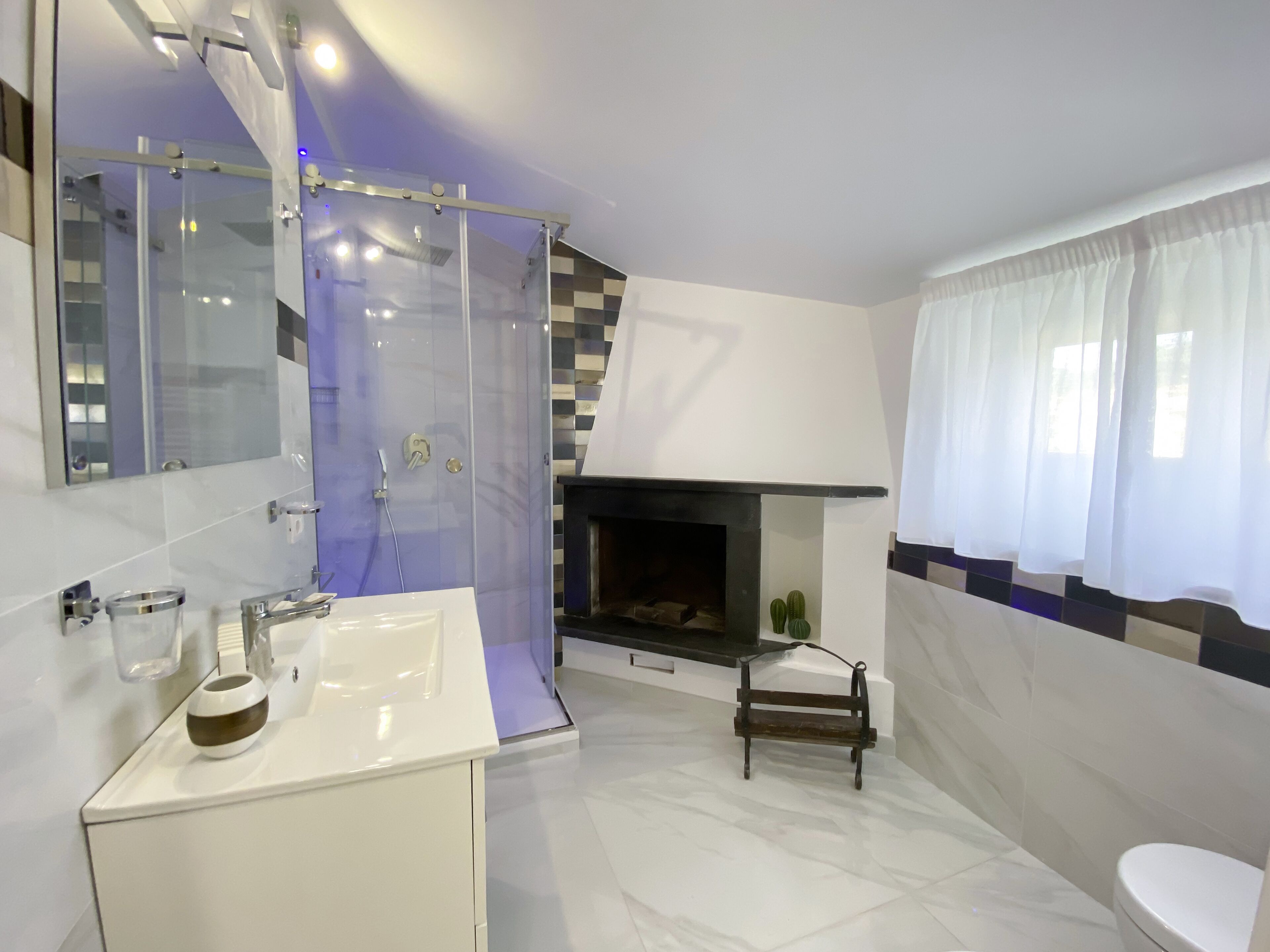 A modern bathroom with a glass shower enclosure, a white sink, a mirror, a fireplace, and a toilet.