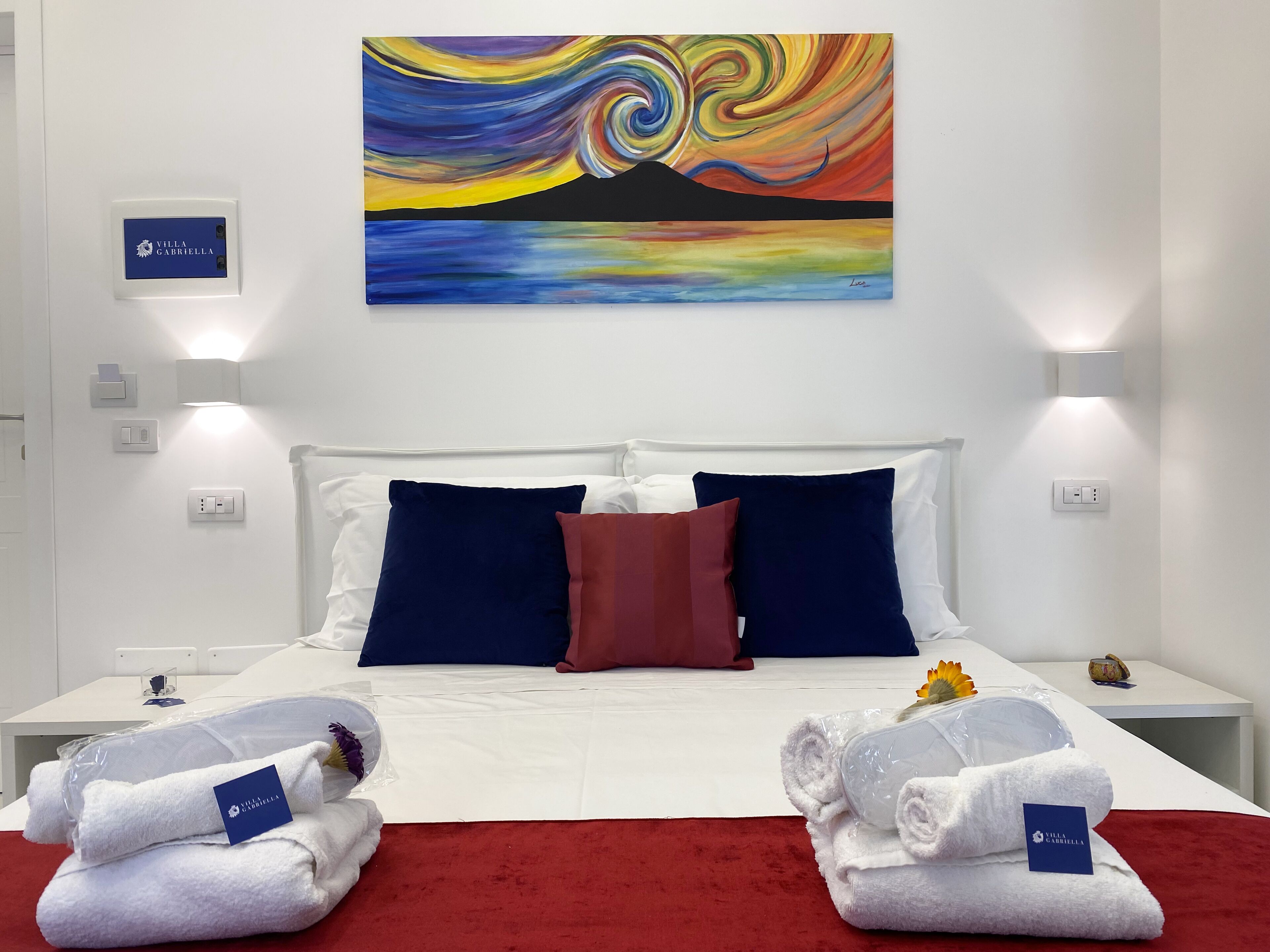 A hotel room with a bed, white walls, a colorful painting, and two white towels with a tag.