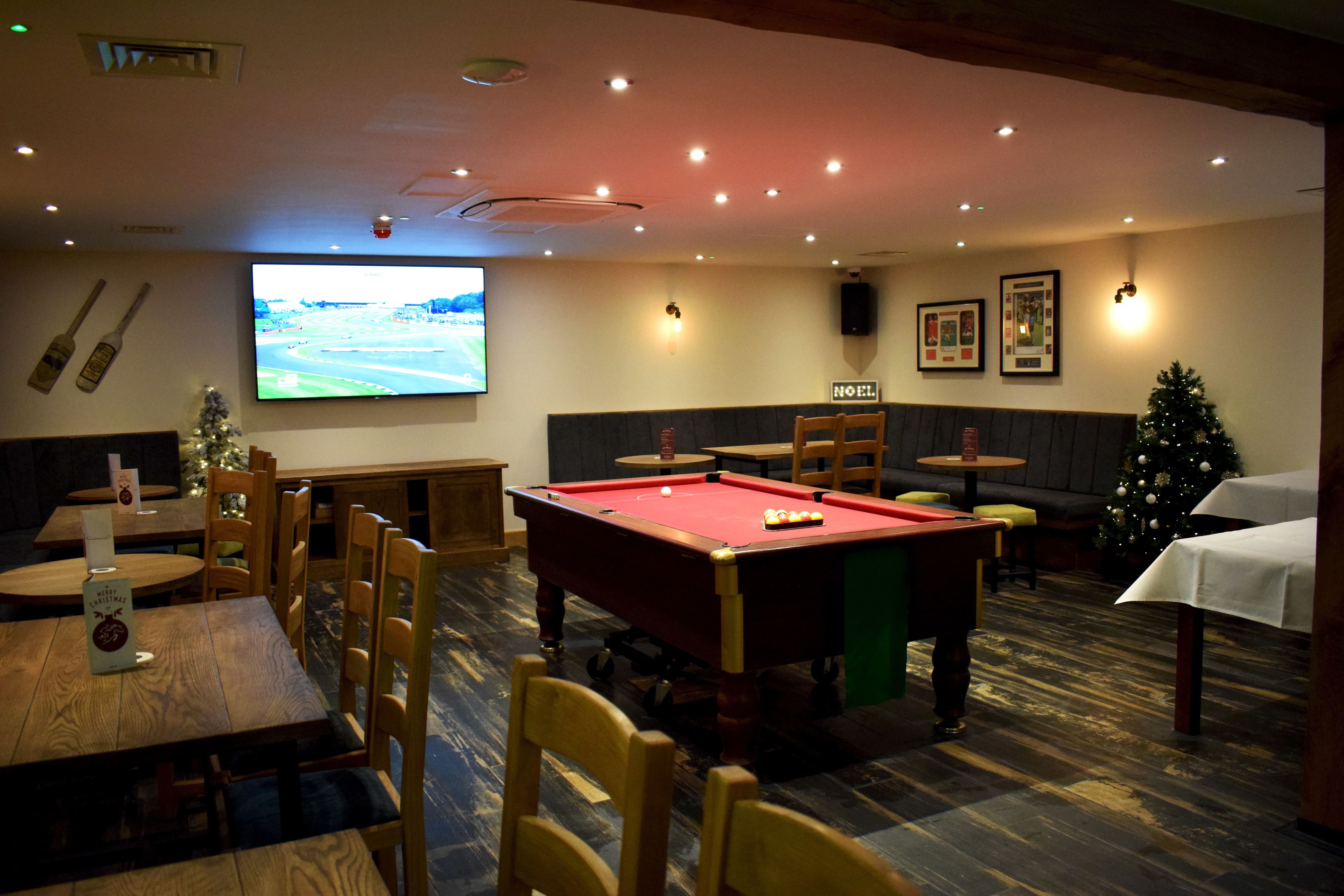Una sala de billar con mesa de pool, mesas y sillas de madera, un árbol de Navidad y una pantalla de televisión.