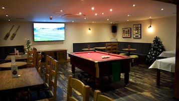 Una sala de billar con mesa de pool, mesas y sillas de madera, un árbol de Navidad y una pantalla de televisión.