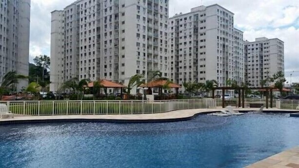 A swimming pool with a surrounding fence, palm trees, and a high-rise building in the background. - Apartamento Barra da Tijuca (Rio de Janeiro)