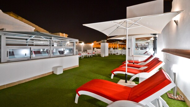 Un bar en la azotea con zona de estar al aire libre, que incluye sillones reclinables rojo y blanco y sombrillas, además de un área de bar con paredes de vidrio.