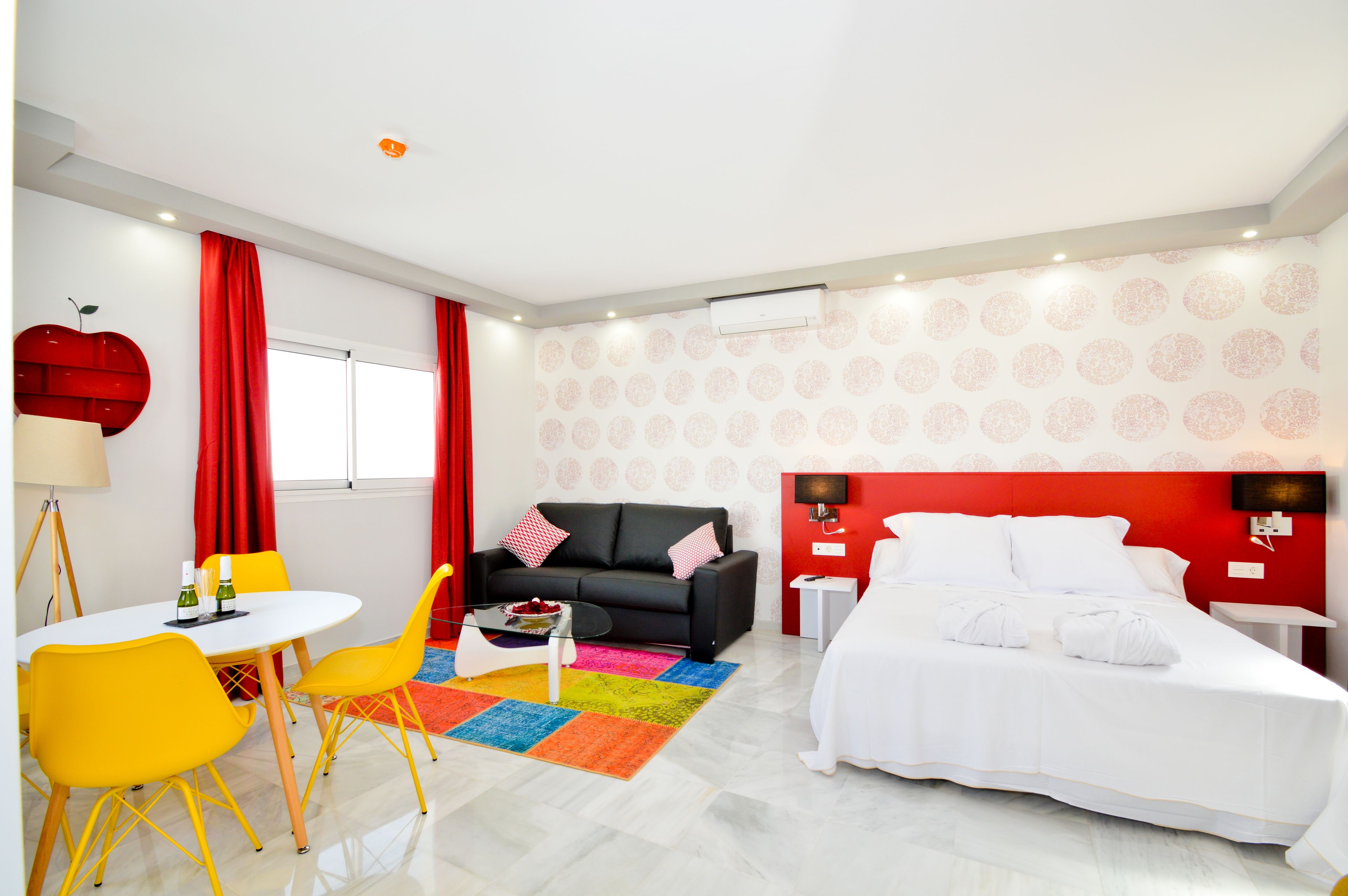 Una habitación de hotel moderna con cama, sofá, mesa de comedor con sillas y una alfombra colorida.