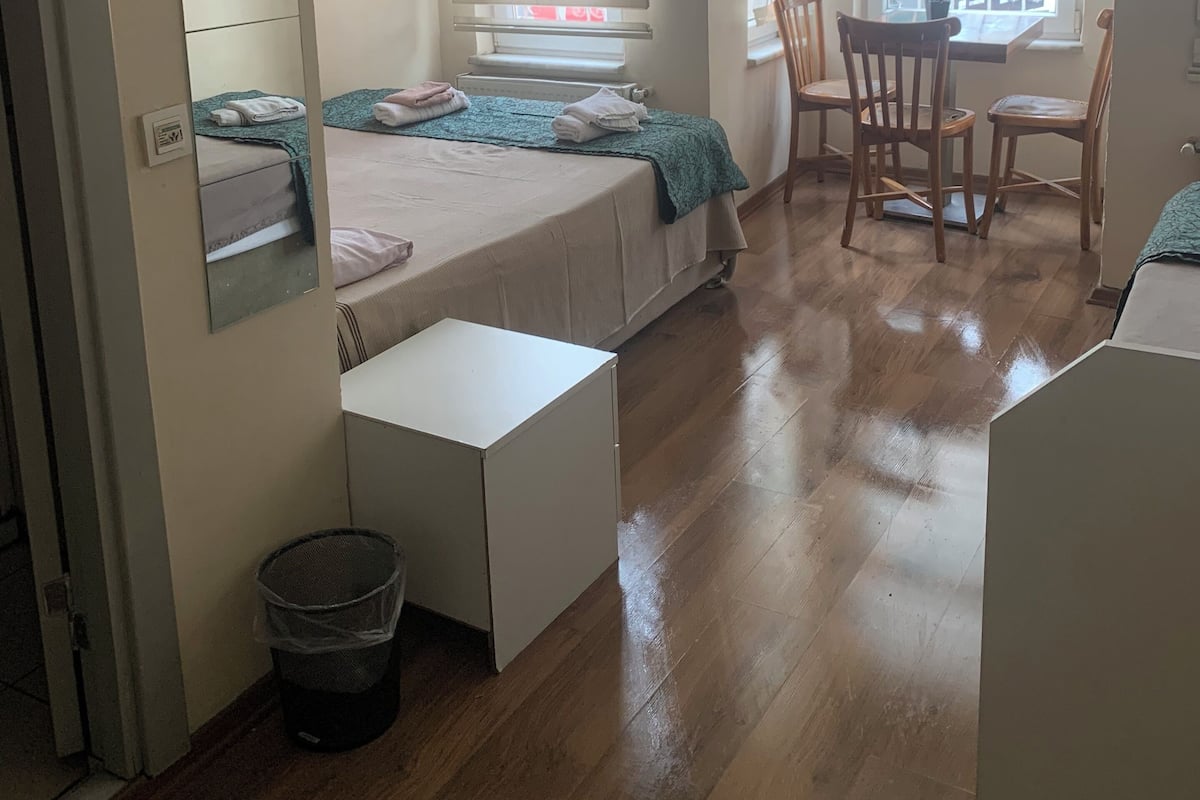 Ein kleines, helles Schlafzimmer mit Bett, Nachttisch und Stuhl. Es gibt ein Fenster mit Jalousien, einen Spiegel und eine kleine Mülltonne.