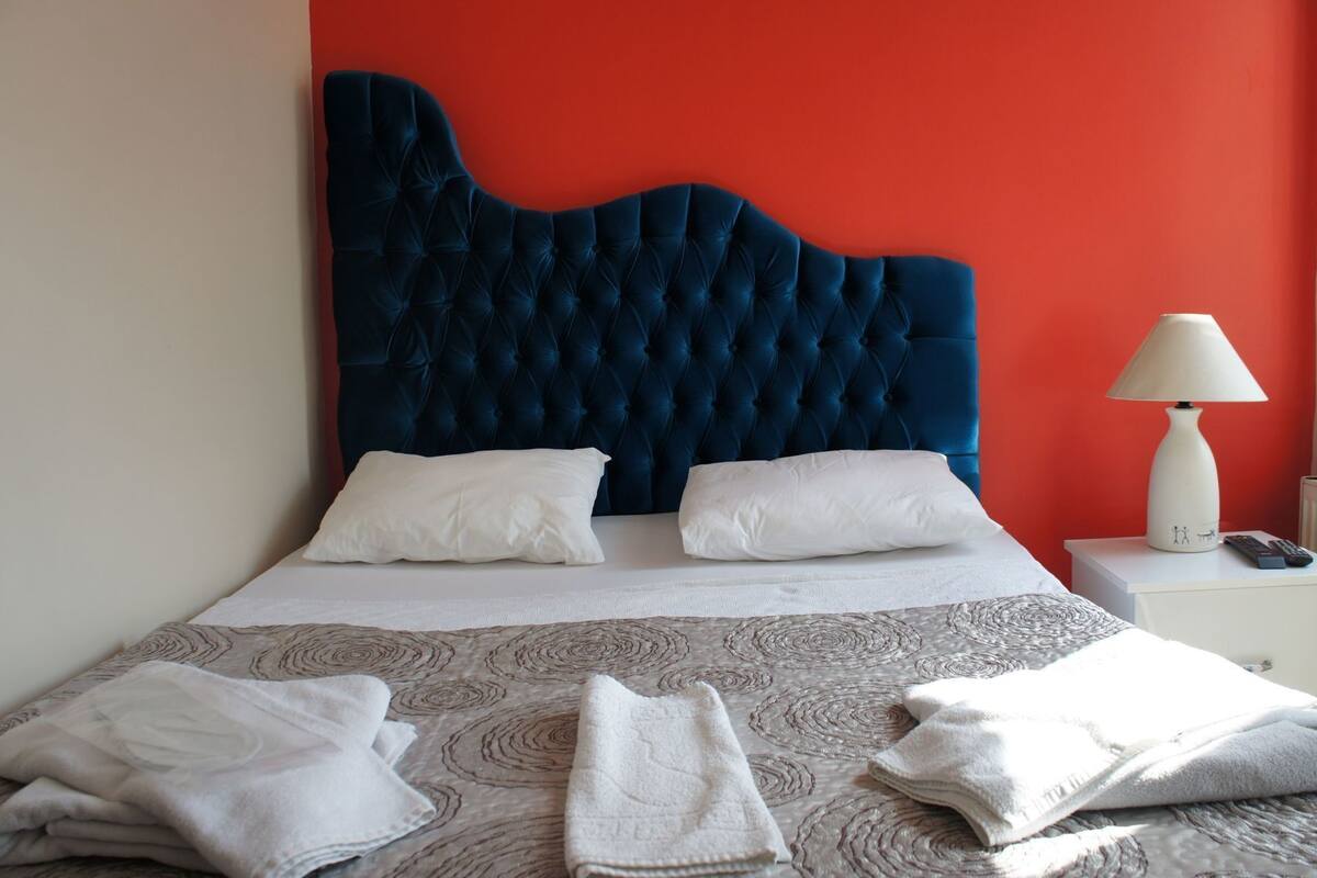 Ein Bett mit einem durchgestopften Kopfteil, weißen Kissen, einem weißen Nachttisch mit Lampe und einer gemusterten Tagesdecke.