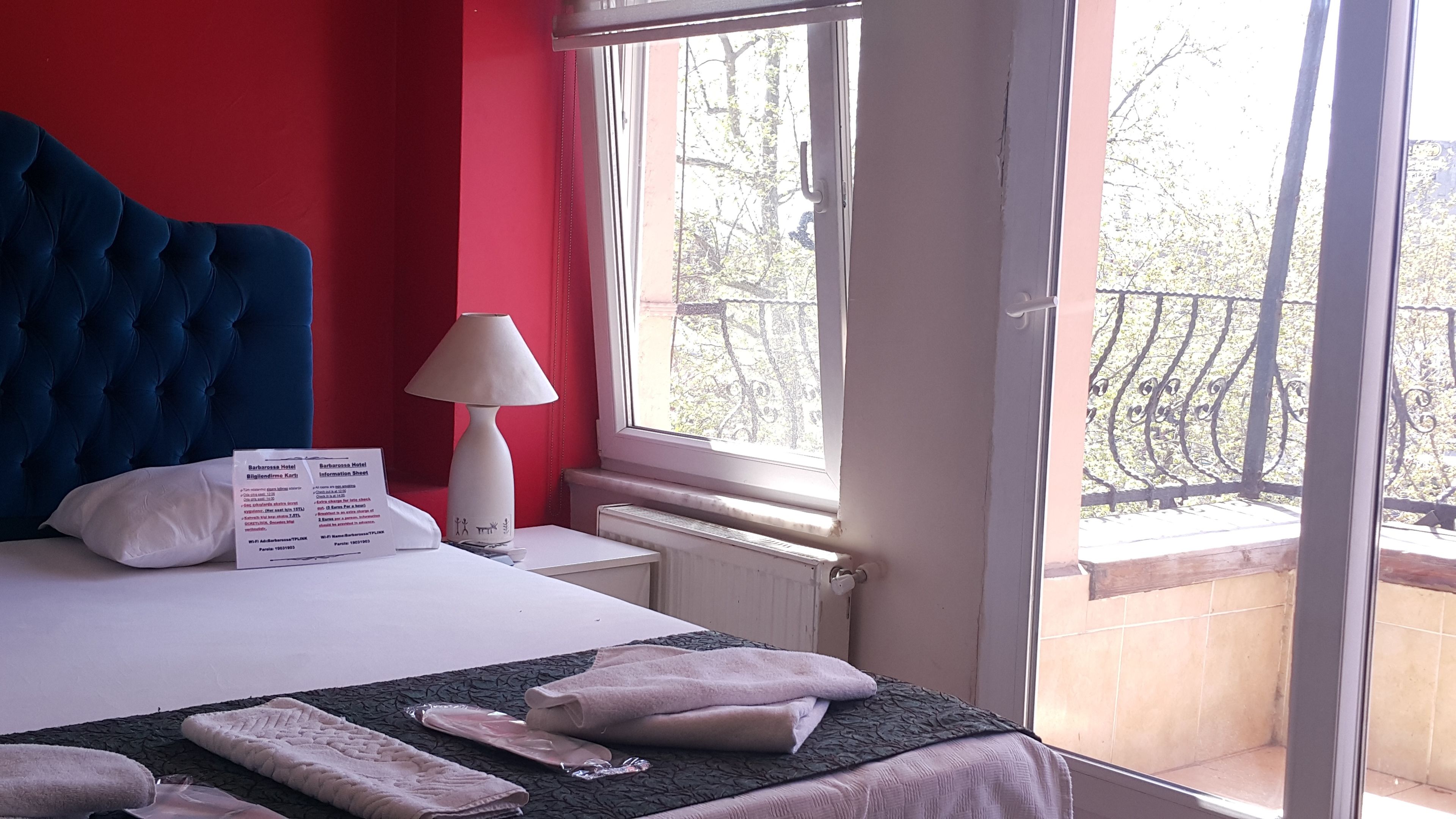 Une chambre d’hôtel avec un grand lit, une table de chevet avec une lampe, une fenêtre donnant sur des arbres et un balcon.