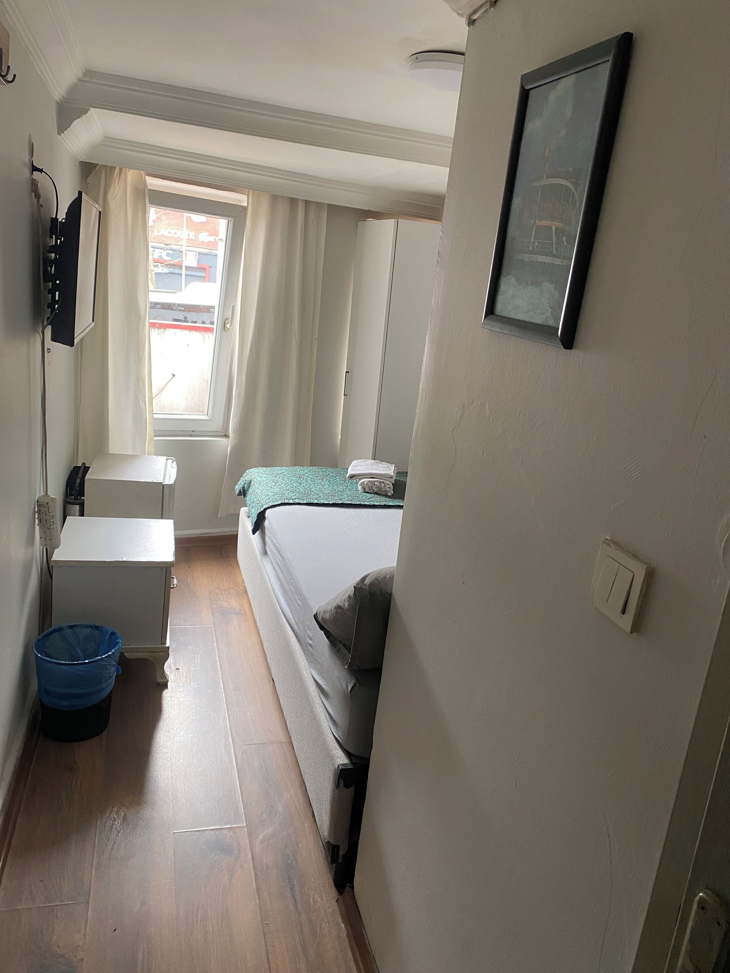 A small, narrow hotel room with a bed, a desk, and a window.