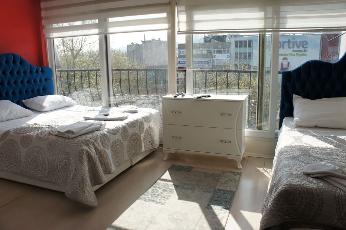 Ein Hotelzimmer mit zwei Betten, einer weißen Kommode und einem großen Fenster mit Blick auf Gebäude und Bäume.